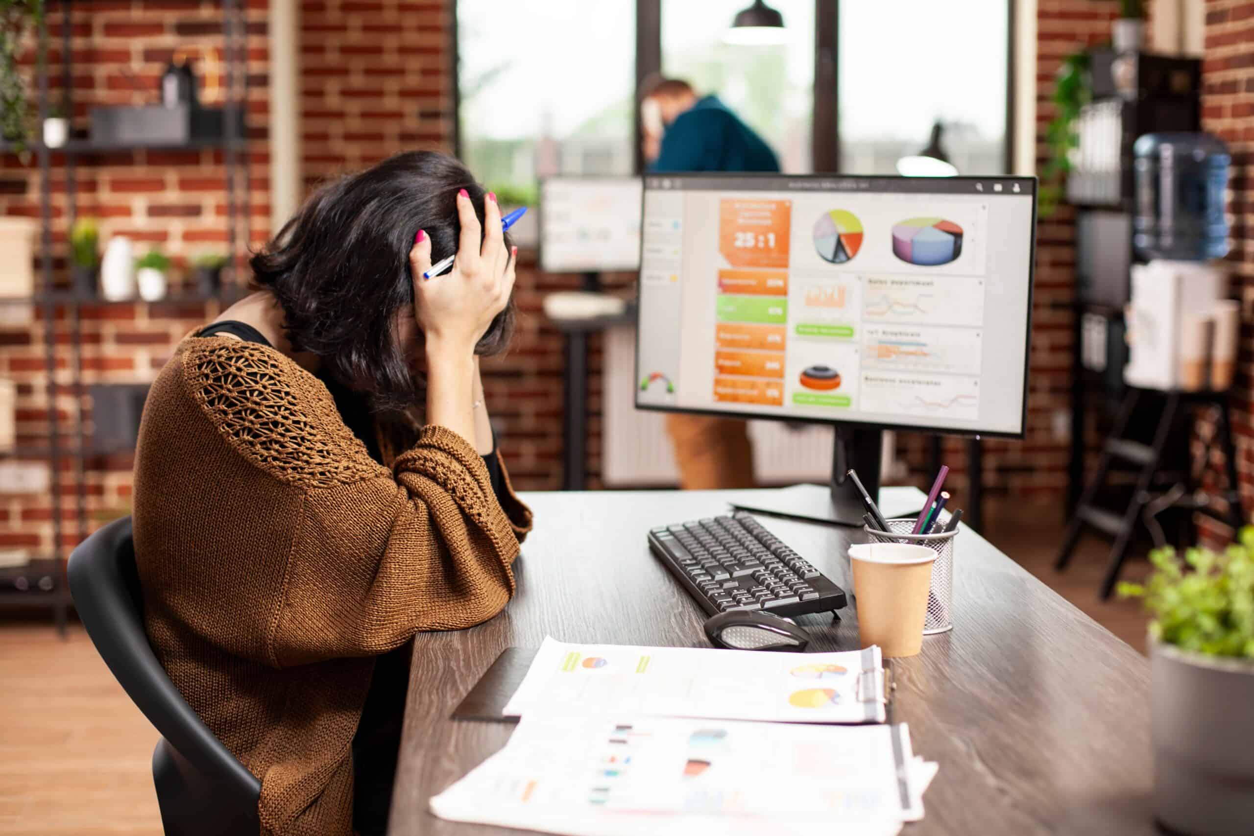 Woman sitting at work desk with computer and head in her hands feeling stress and burnout.
